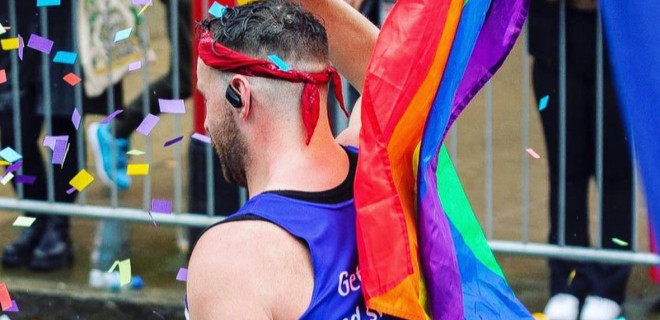 Side shot of a Terrence Higgins Trust marathon runner with the Pride flag in view.