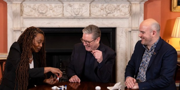 Beverley Knight, Keir Starmer and Richard Angell sit at a table in front of fireplace, performing an HIV test.