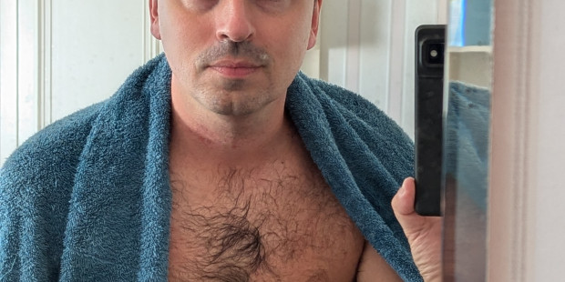 Alex standing with his chest and stomach exposed, draped with a towel and holding a razor.