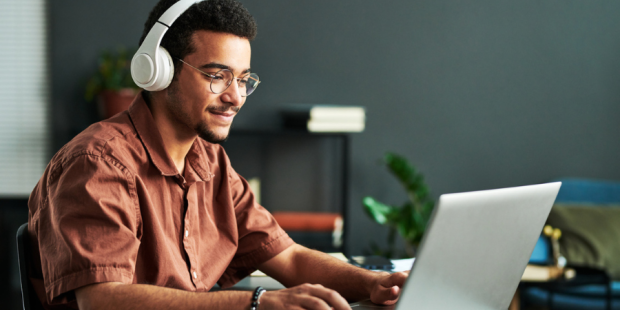 A man sits at a desk wearing headphones and using a laptop.