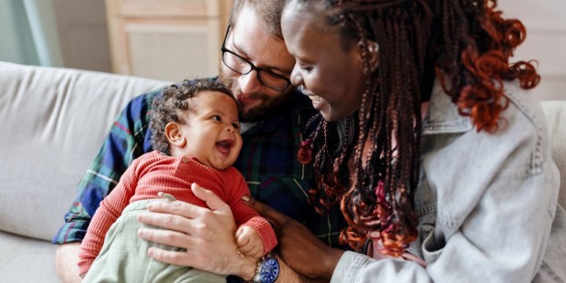 A couple sit on the sofa holding a laughing baby.