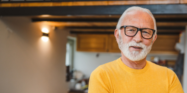 An older man wearing a yellow jumper looks at the camera.